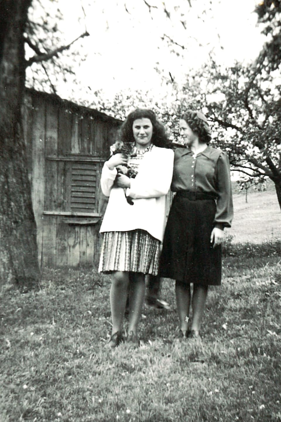 Zwei junge Damen stehen vor Holzhütte. Dorli hält eine Katze auf der Schulter. 