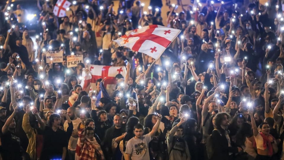 Menschenmenge bei Nacht mit erhobenen leuchtenden Handys und einer Flagge.