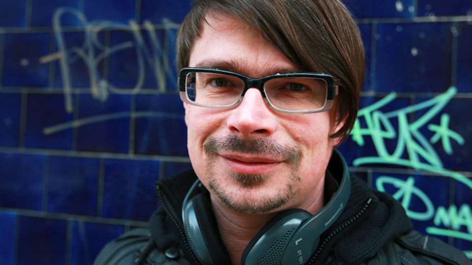 Jaroslav Rudis trägt Kopfhörer um den Hals und eine Brille. Hinten ist eine Wand mit Graffitis.