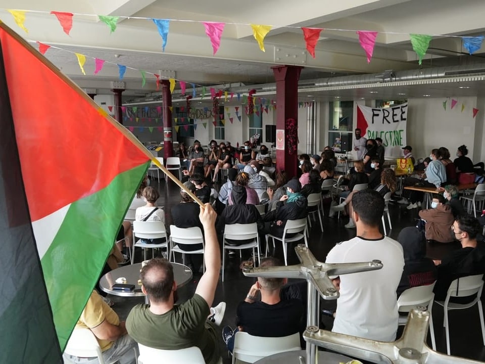 Versammlung in der Mensa mit einer Palästinenser-Flagge, Menschen sitzen auf Stühlen und hören einem Redner zu.