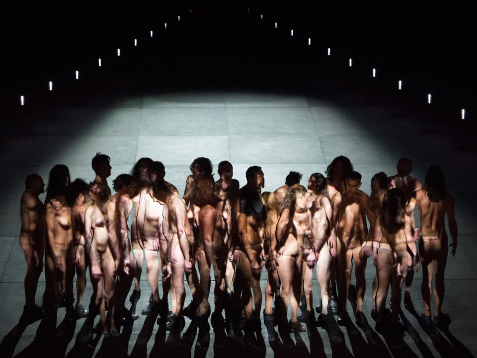 Nackte Männer und Frauen stehen auf einem grauen Bühnenboden.