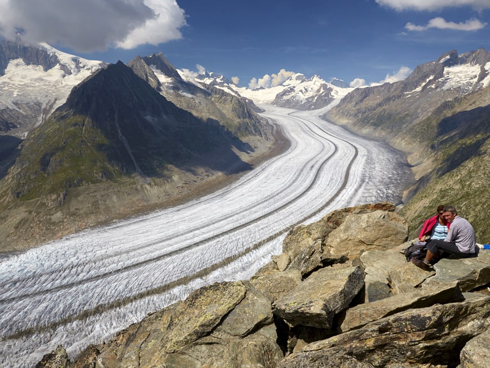 Aletschletscher unten im Tal, zwei Wanderer sitzen auf einem Stein oben.