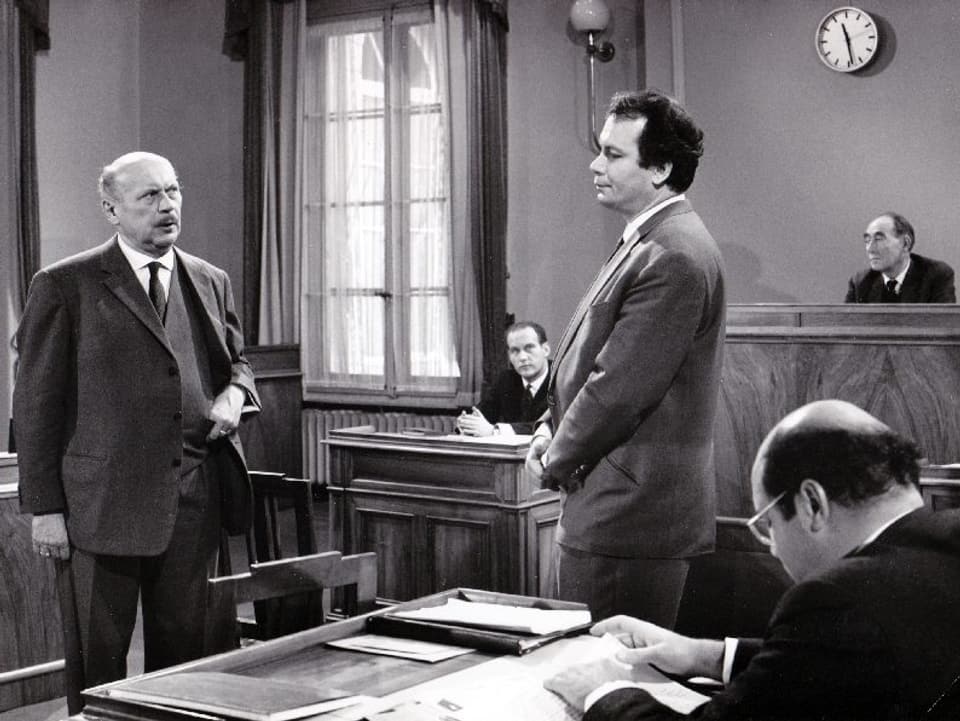 Zwei Männer stehen in einem Gerichtssaal und diskutieren. Drei weitere Männer sitzen auf ihren Plätzen und hören zu.