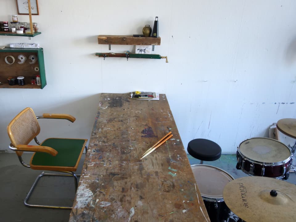 Neben einer grossen Tischplatte mit Spuren von Farbe, stehen verschiedene Trommeln und ein Becken, auf dem Tisch liegen zwei Trommelstöcke. 