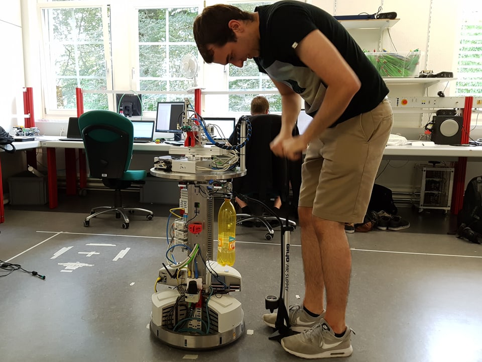 Ein junger Mann pumpt mit einer Velopumpe eine Petflasche auf, die am Roboter montiert ist.