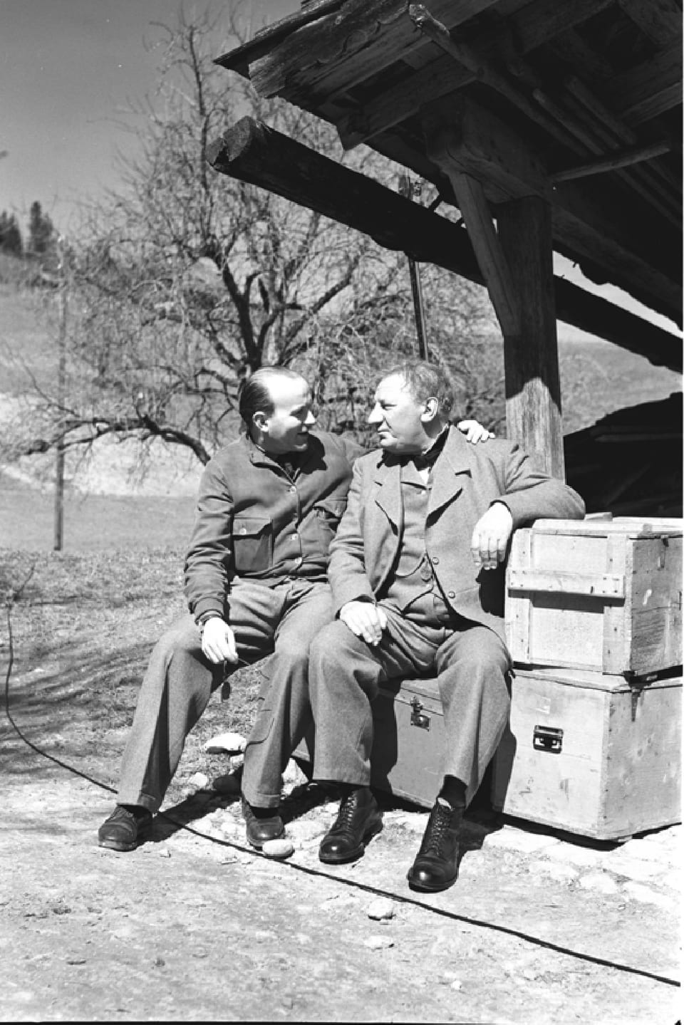 Zwei Männer sitzen auf einer Holzkiste vor einem Schopf und unterhalten sich.