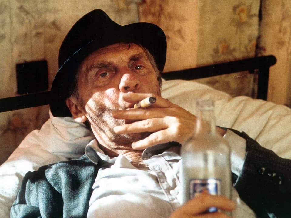 Mann liegt auf Bett mit Flasche Whisky in der Hand und Zigarette im Mund. 