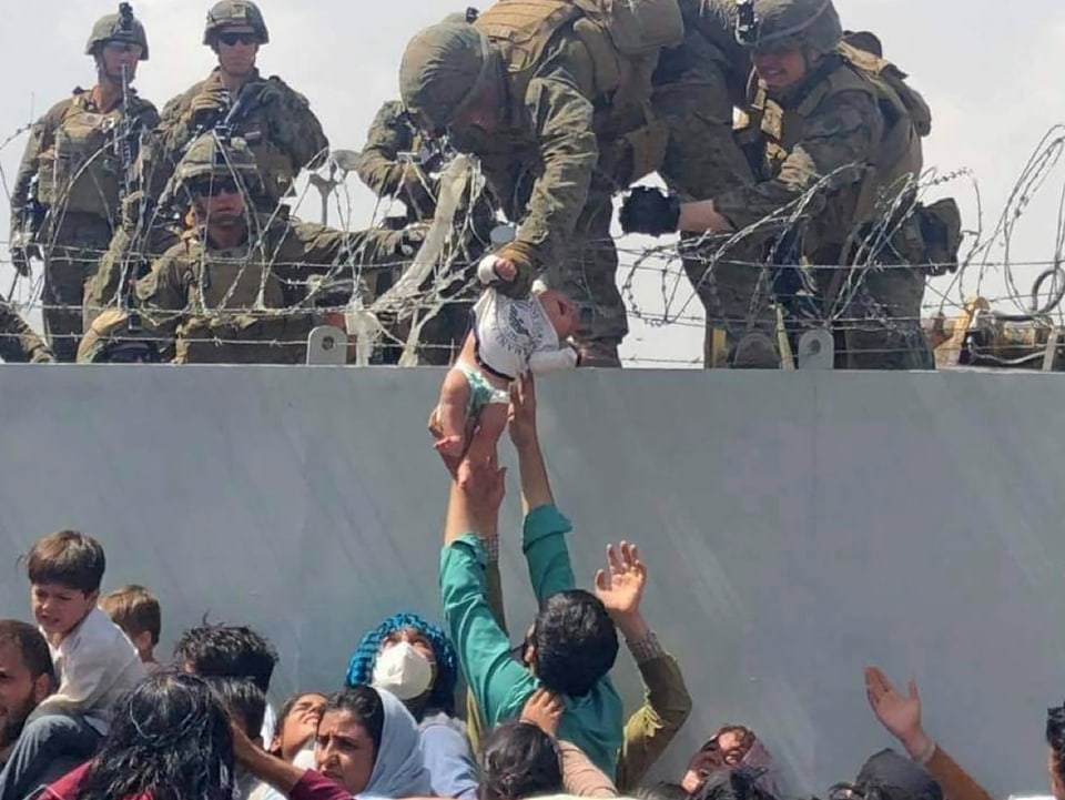 Eine mit Stacheldraht befestigte Mauer trennen Soldaten und Menschen. Ein Mann hält ein Baby nach oben, das von einem Soldaten am Arm gegriffen wird, um es über den Stacheldraht auf seine Seite zu heben. 