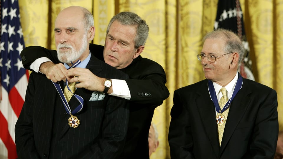 George W. Bush legt Vint Cerf von hinten eine Medaille um den Hals, Bob Kahn schaut rechts davon stehend skeptisch zu.