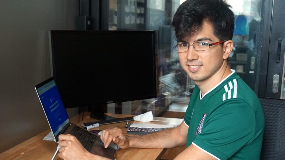 Ein junger Mann mit einem Fussballtrikot, dunklen Haaren und einer Brille sitzt vor einem Laptop.