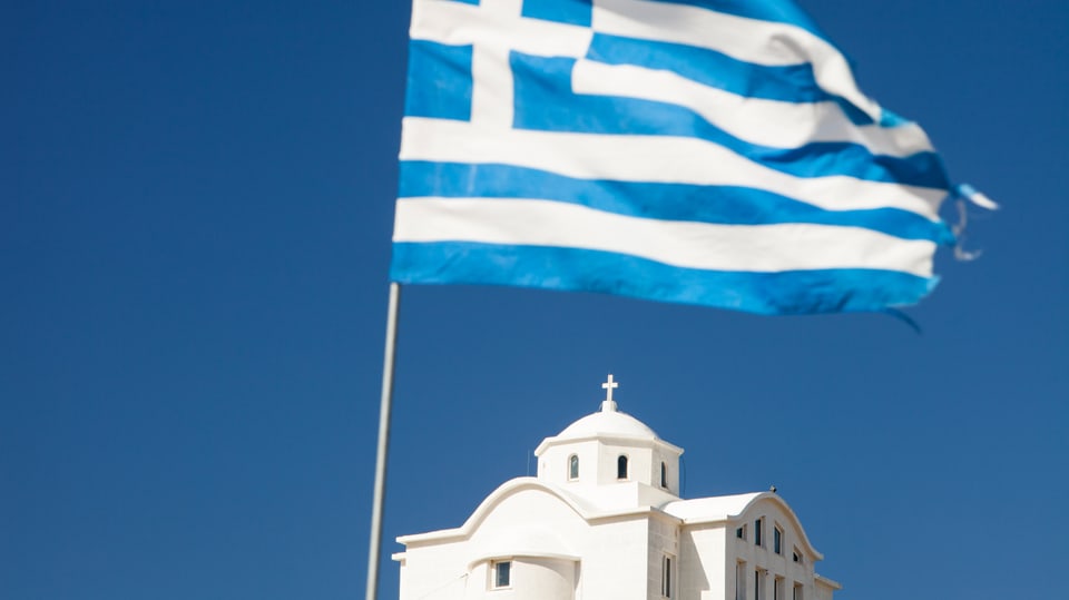 Eine griechische Flagge weht vor einer orthodoxen Kirche im Hintergrund.