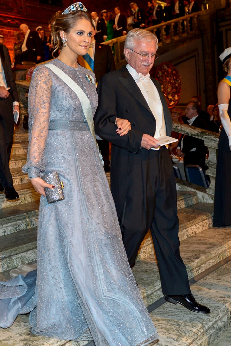 Prinzessin Madeleine in einem blau-grauen Abendkleid mit einem Mann am Arm eine Treppe hinunterschreitend.