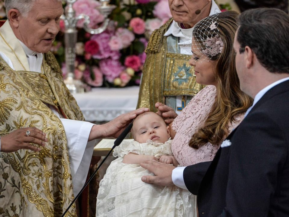 Prinzessin Madeleine hält Leonore auf dem Arm. Diese hat die Augen zu während der Pfarrer ihr den Kopf berührt. Chris O'Neill berührt Leonore mit einer Hand.
