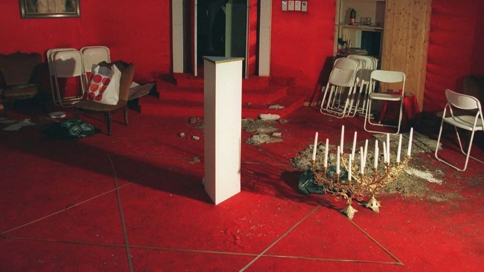 Raum mit rotem Teppich, ein Stern darauf gezeichnet, ein Kronleuchter und Asche am Boden.