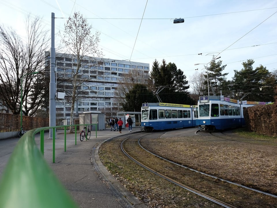 Zwei blau-weisse Trams stehen an der Endstation, deren Wendeplatz mit einem grünen Geländer umgeben ist.