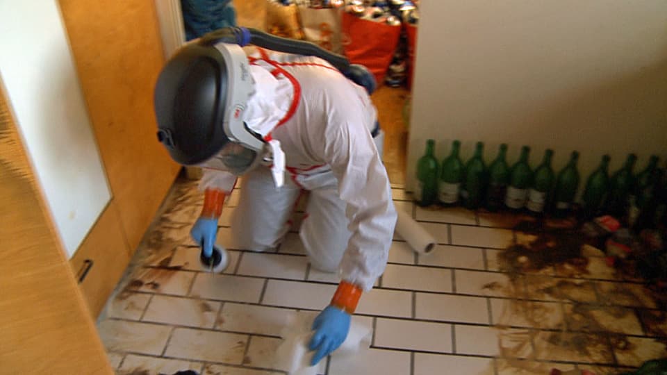 Ein Mann mit Atemmaske und Schutzanzug putzt den Boden in einer Wohnung.
