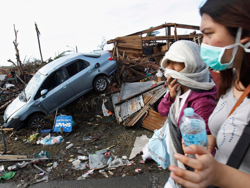 Eine Frau mit einem Baby auf dem Arm, eine Atemmaske tragend, steht vor den Trümmern ihres Hauses.