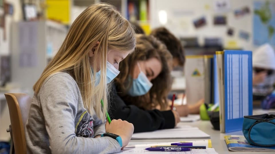 Ein Schulkind sitzt mit Maske an einem Tisch in der Primarschule und löst Aufgaben.