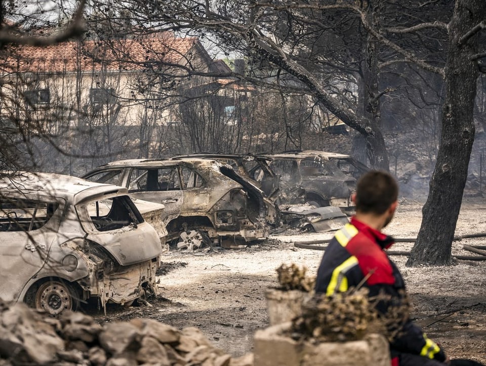 Ein Feuerwehrmann sitz im Vordergrund des Bild. Im Hintergrund sind ausgebrannte Autos und Bäume zu sehen.