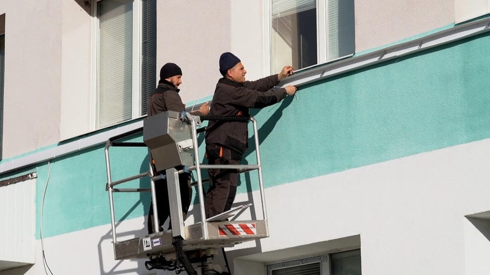 Zwei Männer stehen auf einem Hebekran an einer Häuserwand und befestigen Leisten für Stromkabel..