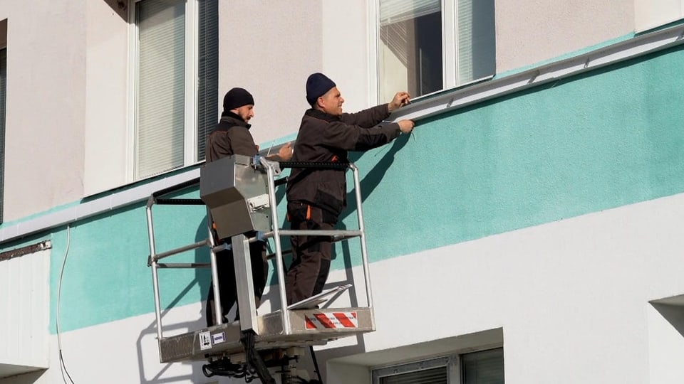 Zwei Männer stehen  auf  einem Hebekran an  einer Häuserwand  und befestigen Leisten für Stromkabel..