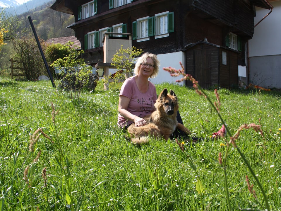Heidy Gasser sitzt mit ihrem Hund auf einer Wiese vor einem dunklen Haus mit grünen Fensterläden.