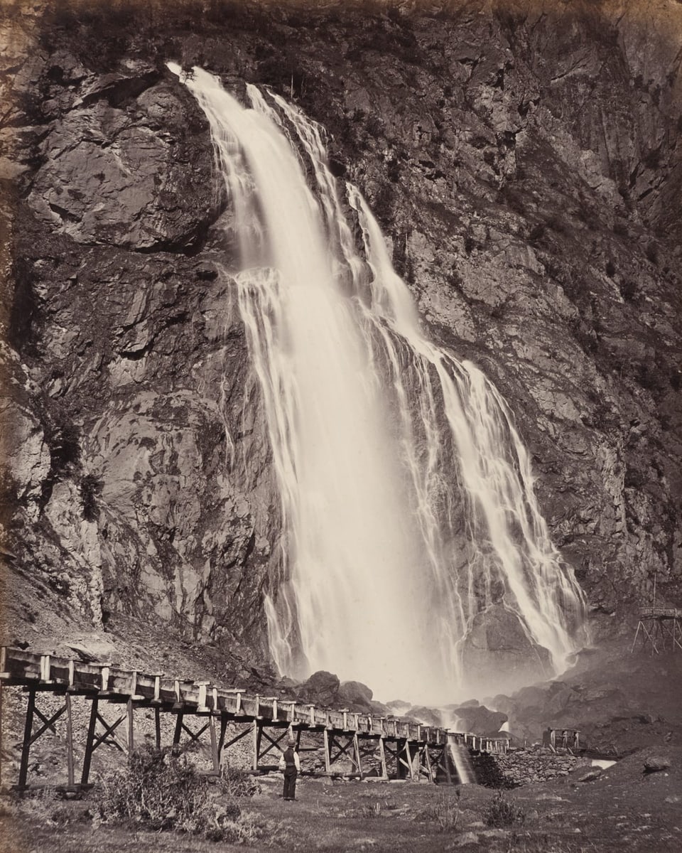 schwarzweiss Fotografie Wasserfall, davor eine Holzkonstellation, in der Wiese steht ein Mann mit Hut