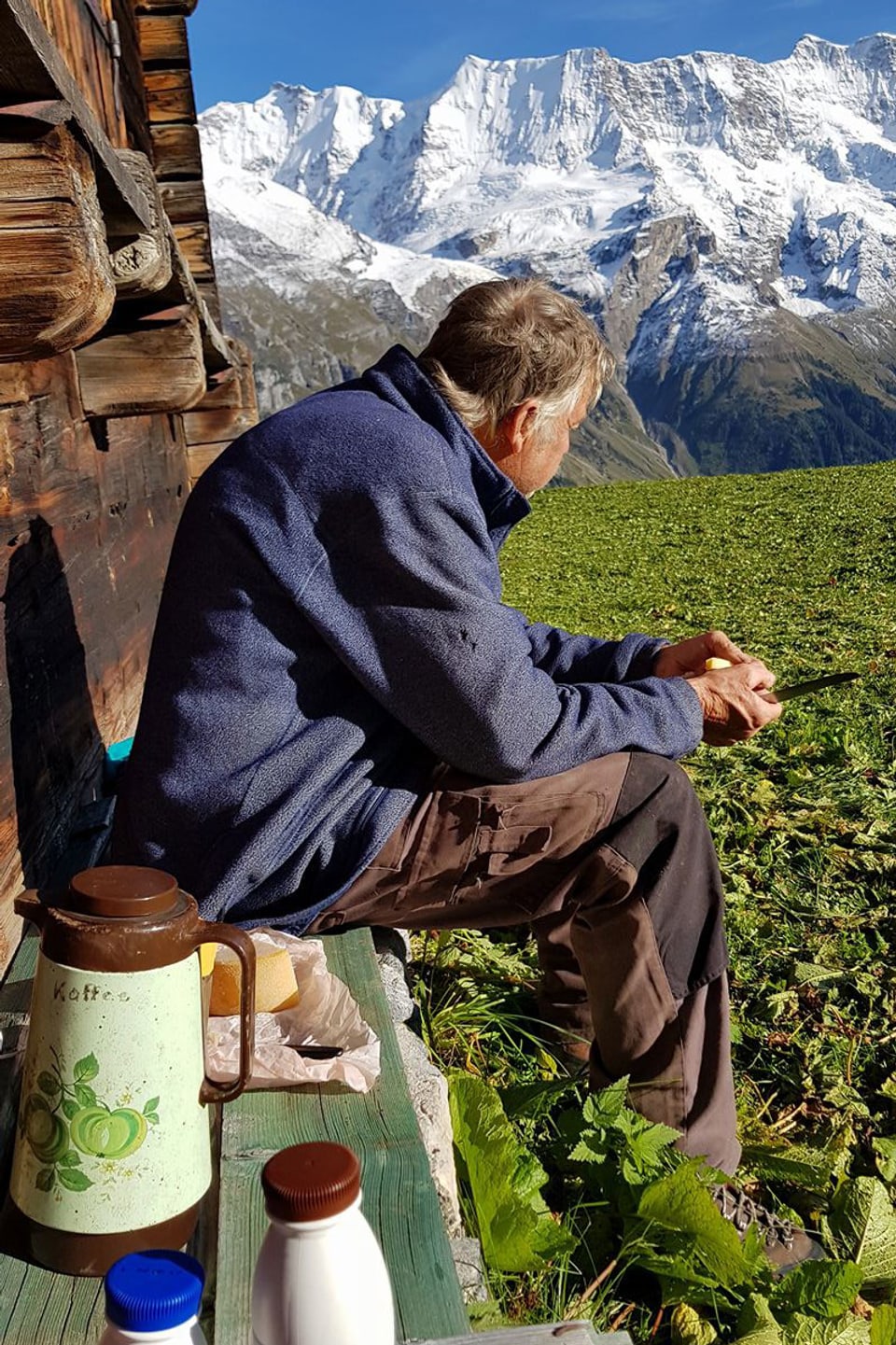 Ein älterer Herr sitzt vor einem alten Holzhaus und isst ein Picknick.