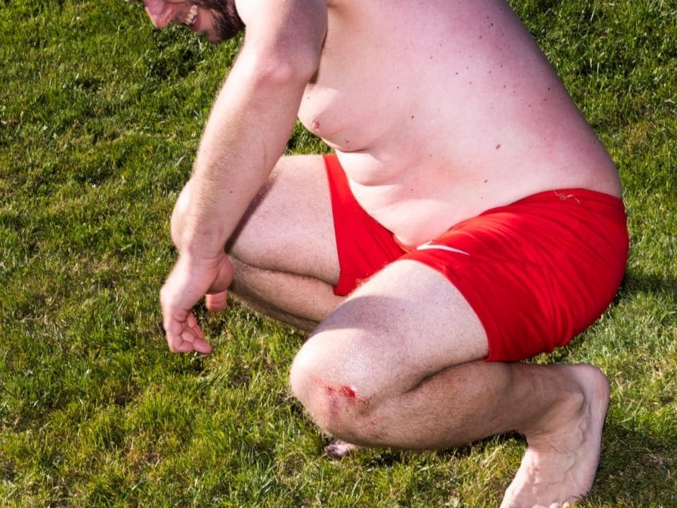 Ein unsportlicher Mann mittleren Alters geht auf einer Wiese in roter Badehose in die Hocke. Er lacht.