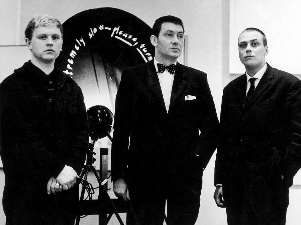 Drei Männer in schwarzen Anzügen, nebeneinander stehend (s/w).