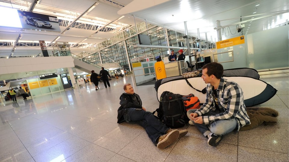 Zwei junge Männer liegen in einer Flughafenhalle am Boden.