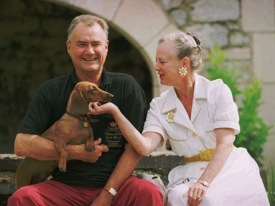 Ein Bild aus den 90ern von Prinz Henrik und Königin Margrethe. Er hält einen Dackel auf dem Arm. Sie streichelt den Hund. Er trägt eine rote Hose und ein schwarzes Poloshirt. Sie ein weisses Kleid und Brille.