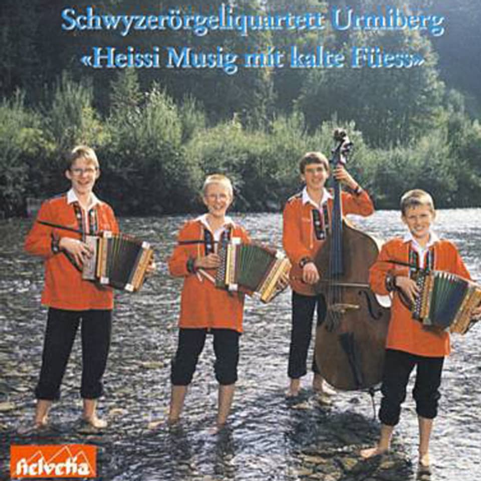Vier Jungs mit nackten Füssen und Instrumenten mitten in einem Fluss. 