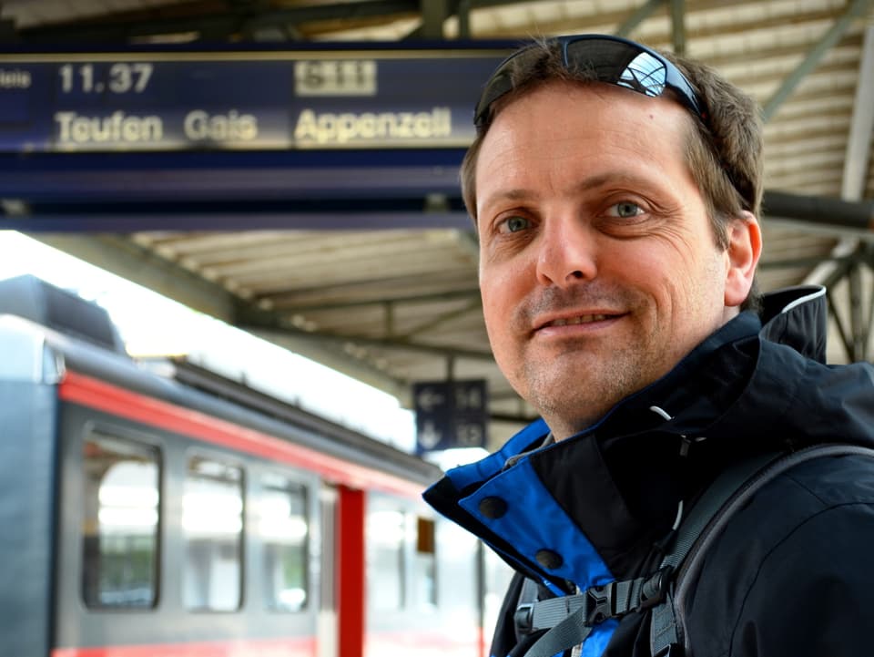 Ein Mann steht an einem Bahnhof. Im Hintergrund die Anzeigetafel mit der Aufschrift "Appenzell".