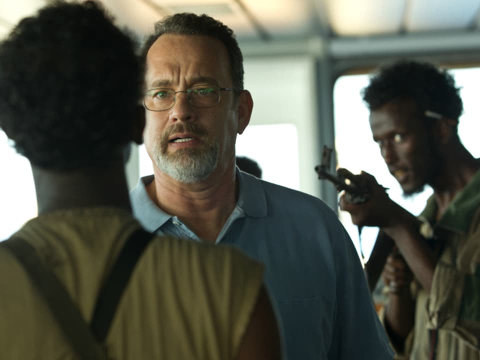 Filmszene: Tom Hanks als Schiffskapitän zwischen zwei Piraten. Einer richtet sein Gewehr auf ihn.