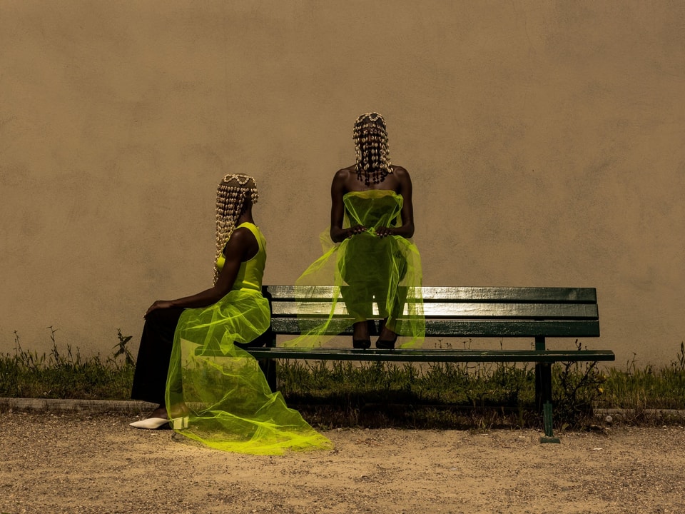 Zwei schwarze Frauen in neongelbem Tüll und mit Holzperlenschmuck sitzen auf einer Bank.
