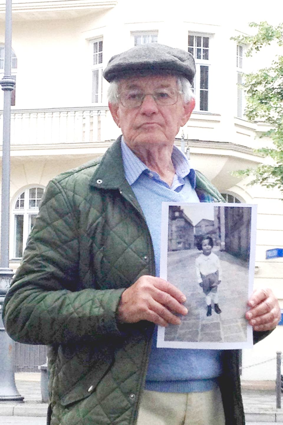 Ein älterer hält ein Foto von sich als Junge in der Hand und steht vor einem historisierenden Haus.