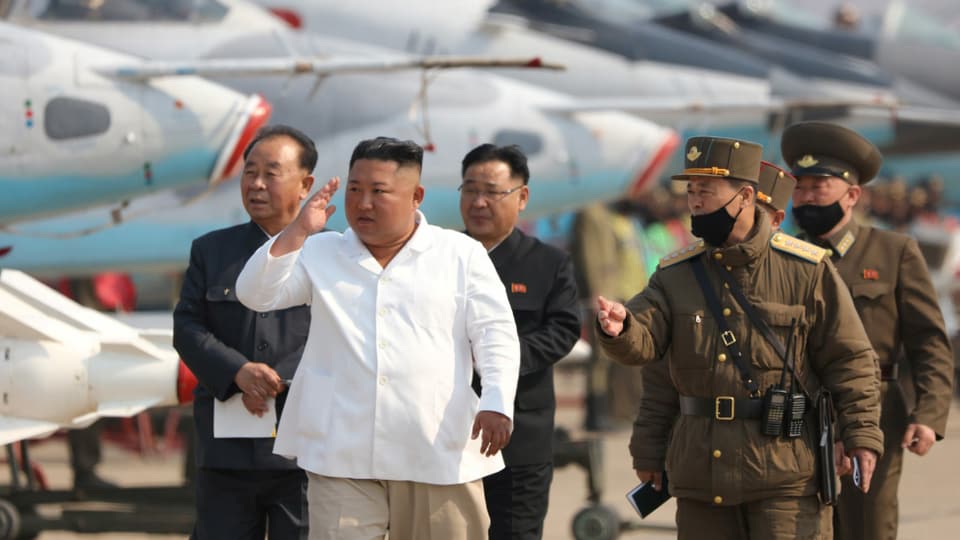Kim in weissem Hemd, umringt von Militärs, einige tragen schwarzen Mund- und Nasenschutz, dahinter aufgereiht Kampfflugzeuge.