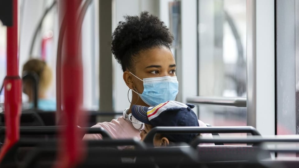 Eine Mutter sitzt im Bus mit ihrem Sohn und zeigt aus dem Fenster, beide tragen Masken.