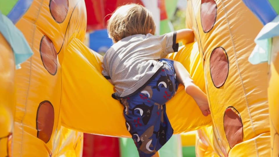 Kind in T-Shirt und Shorts (von hinten) klettert auf einer gelben Hüpfburg mit braunen Punkten. 