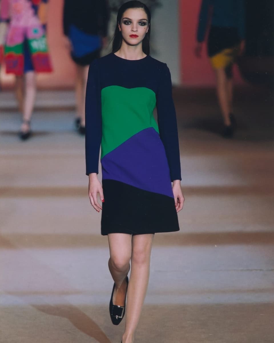Woman on catwalk, hair slicked back, black dress with green and blue shapes.