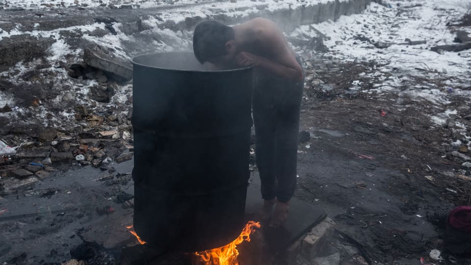 Ein Junge beugt sich in eine Abfalltonne, unter der ein Feuer brennt. Er trägt nur eine Hose und es liegt Schnee.