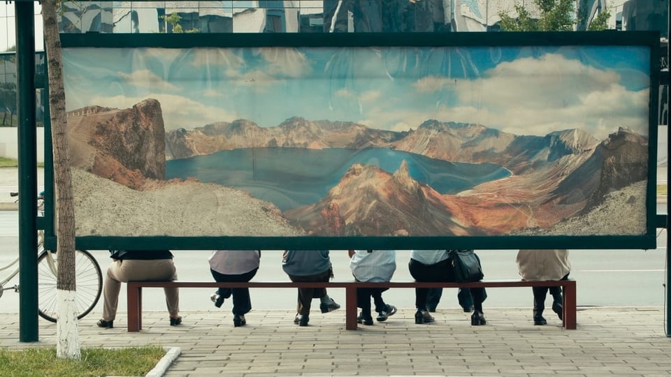grosses Bergplakat verdeckt Bushaltestelle, darunter schauen Beine von Leuten auf einer Bank hervor.
