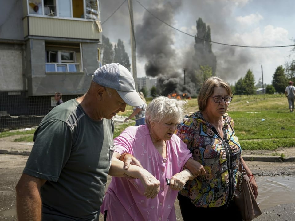 Drei Personen vor einem rauchenden Haus nach Beschuss.