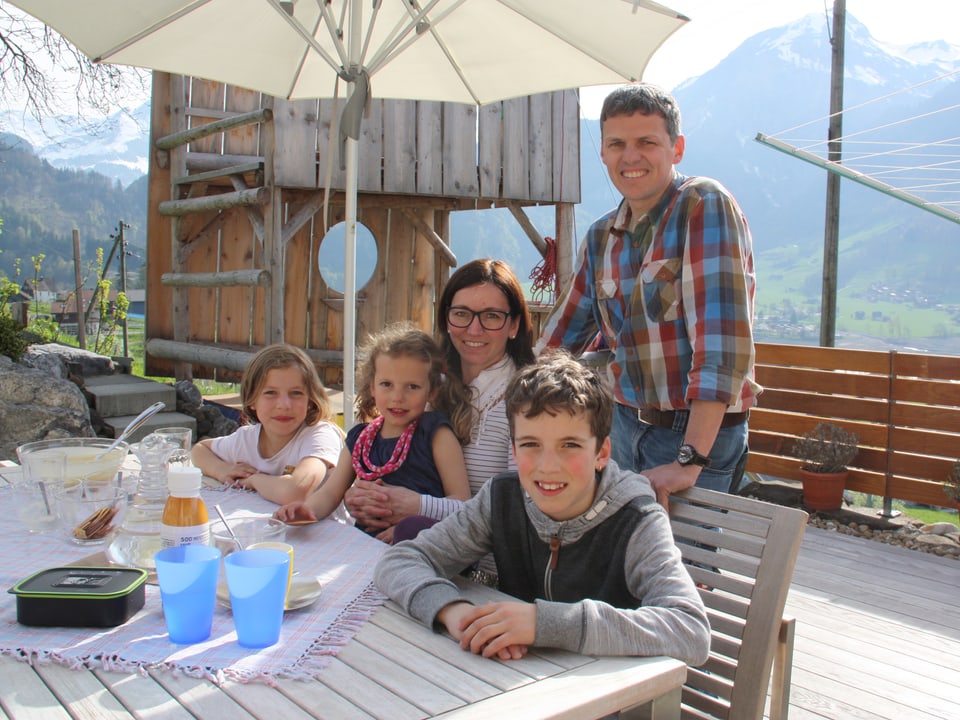 Eine fünfköpfige Familie sitzt auf einer Terrasse an einem Holztisch.