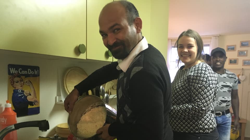 Ein Mann mit einer Pfanne voll Reis steht in einer Küche, daneben eine Frau.