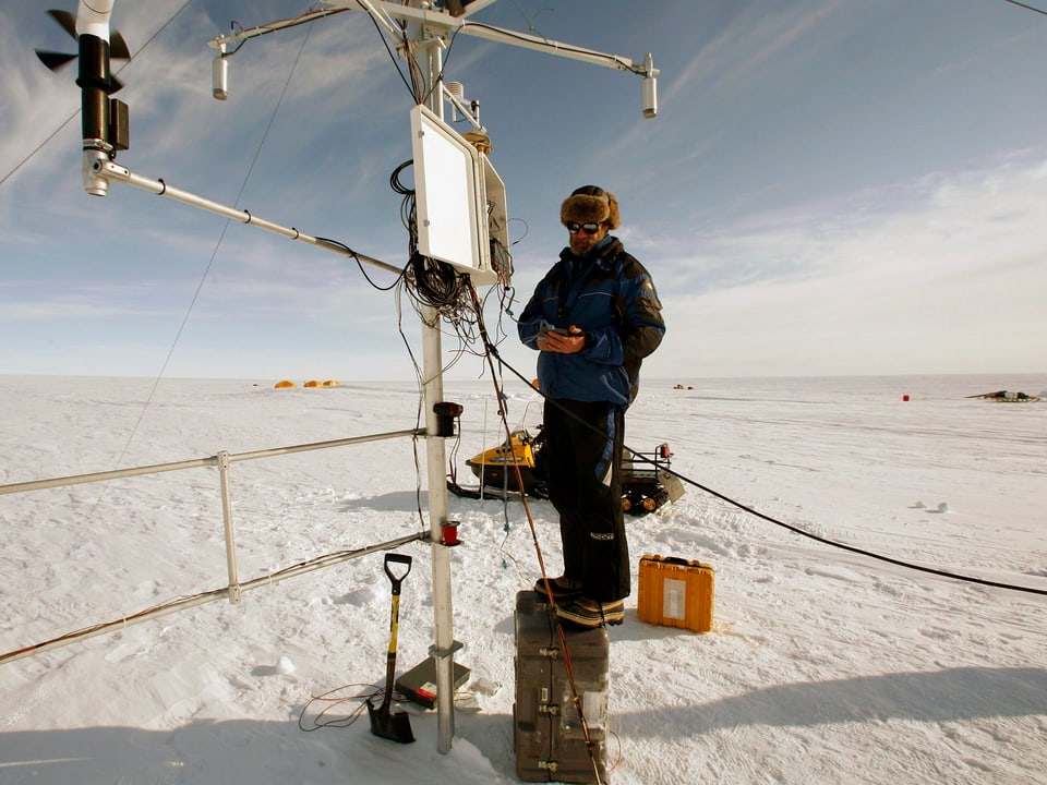 Ein Forscher steht in Winterkleider verpackt auf einer Kiste und wartet die Wetterstation auf dem Gletscher. Ausser ein paar Zelte ist sonst nichts als eine weisse, flache Landschaft zu sehen.