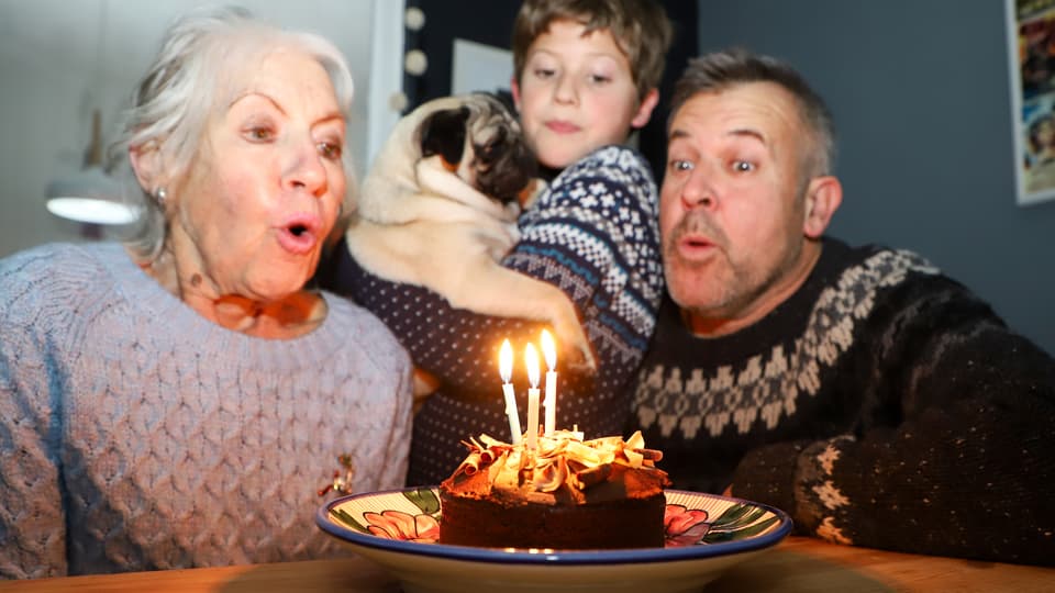 Eine ältere Frau sitzt mit einem Mann und einem Kind an einem Tisch und bläst drei Kerzen auf einer Torte aus.