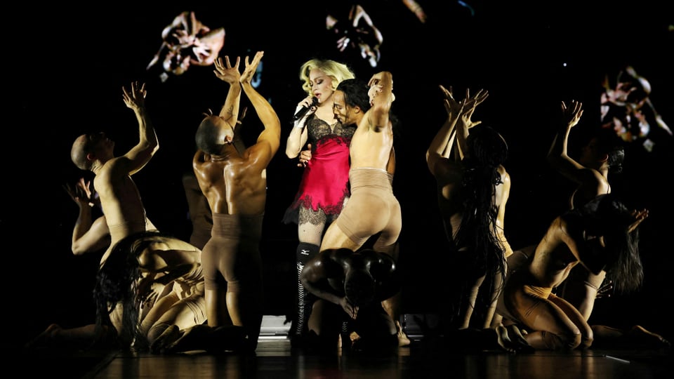 Madonna wears a red and black nightgown, surrounded by people wearing skin-colored suits. 