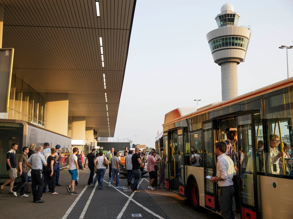 Personen steigen in einen Bus ein. Im Hintergrund ist der Funkturm eines Flughafens zu sehen.